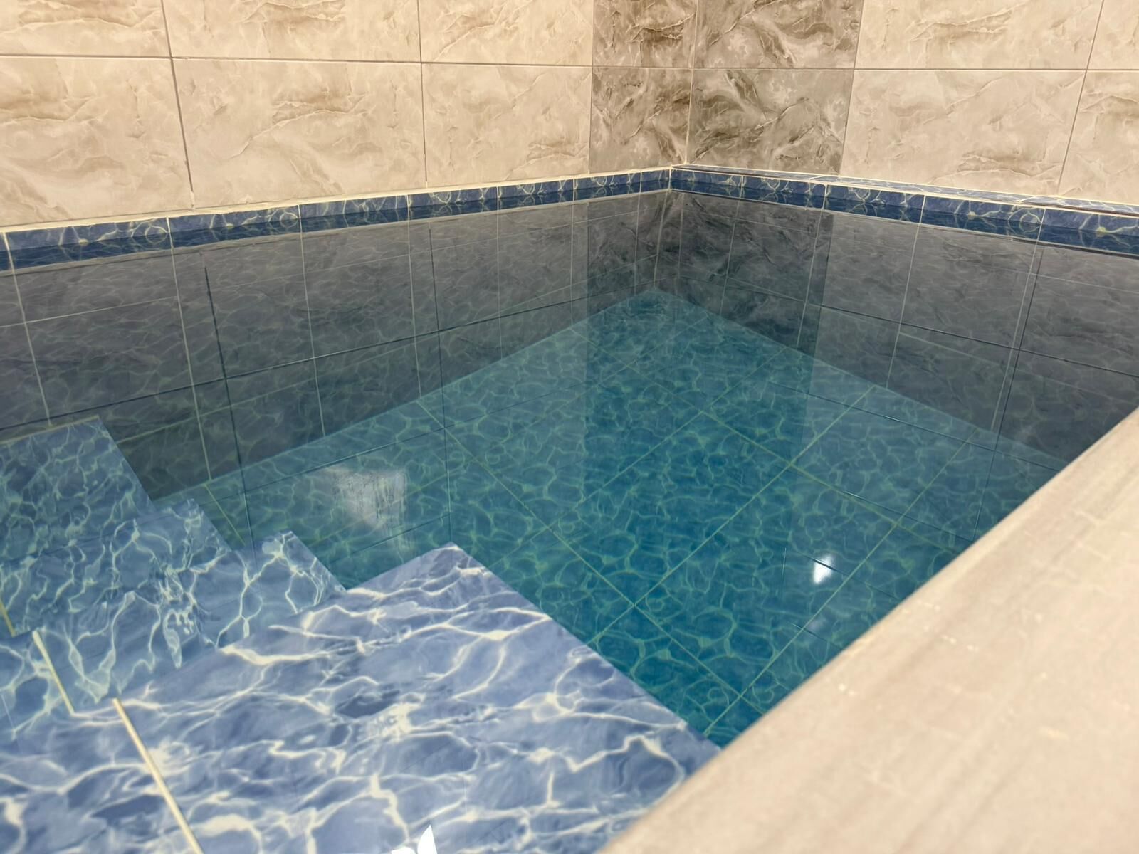 A clear pool with a blue marble pattern on the floor, surrounded by beige tiles.