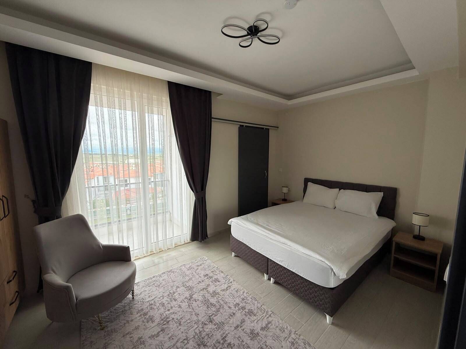 A hotel room with a bed, a chair, a nightstand, a lamp, and a view of the outside.