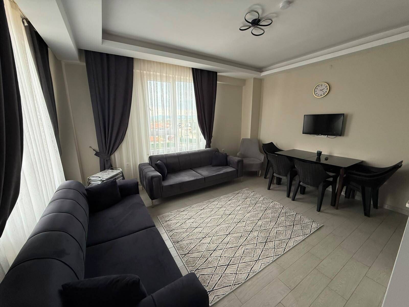 Ein modernes Wohnzimmer mit einer Couch, einem Esstisch mit Stühlen, einem an der Wand befestigten Fernseher und einem gemusterten Teppich.