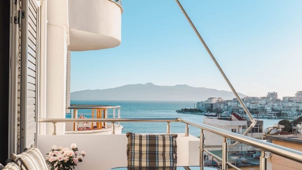 A balcony with a table and chairs, a potted plant, and a view of the city and sea. - Albania Dream Saranda Apartment (Sarandë)