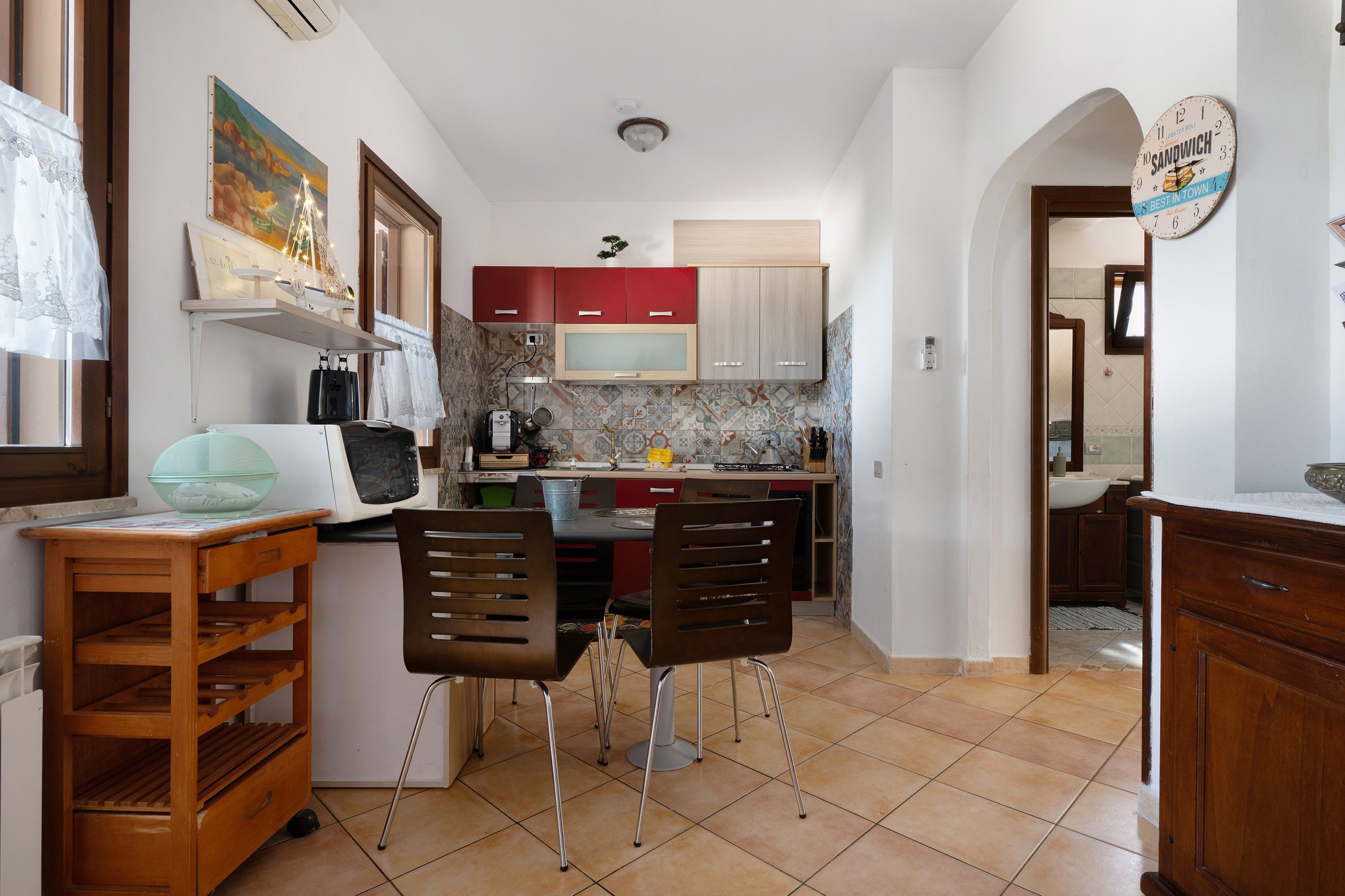 Cocina compacta con microondas, estufa y armarios rojos. Hay un comedor con dos sillas y una mesita.