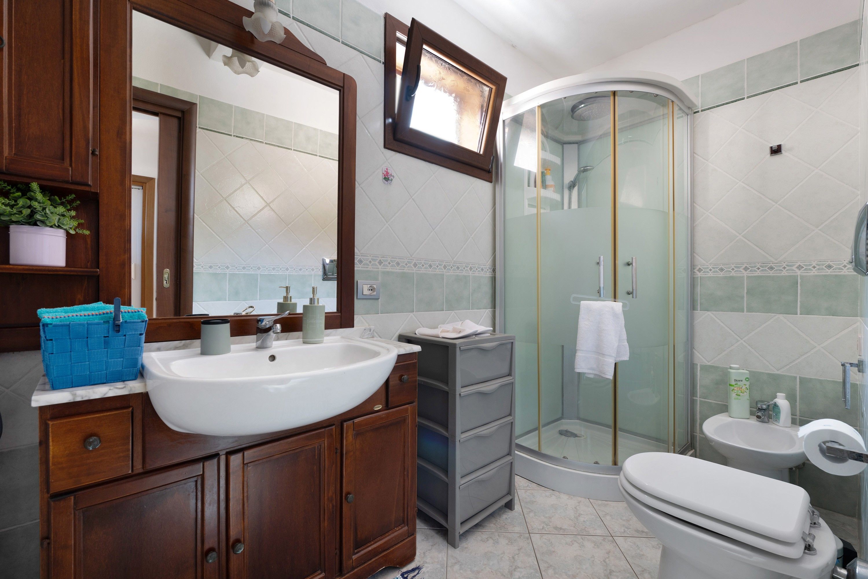 Ein Badezimmer mit einem Holzkosmetikschrank, einem großen Spiegel, einem weißen Waschbecken, einer Glasdusche und einer Toilette.