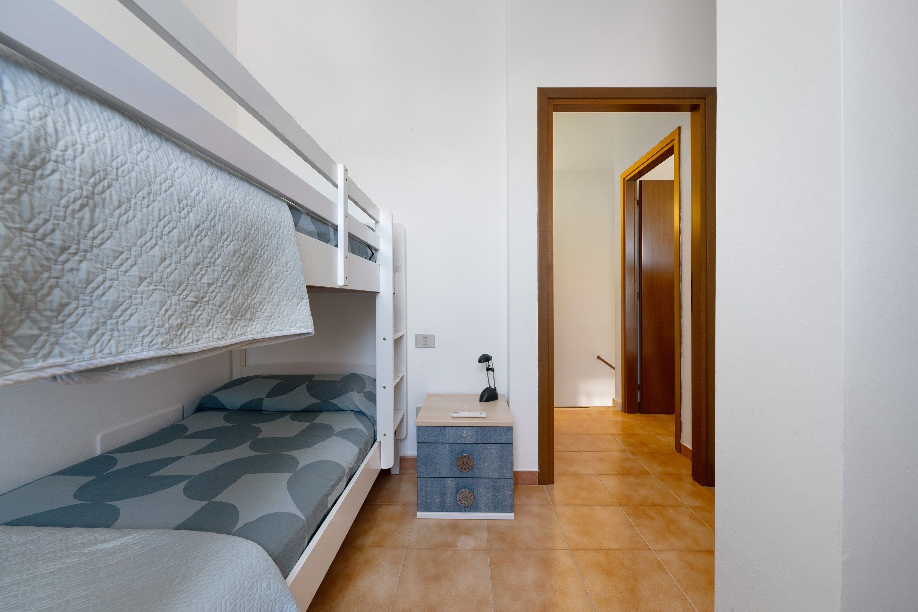 Una stanza compatta con un letto a castello, un comodino con una lampada e una porta che dà su un'altra stanza.