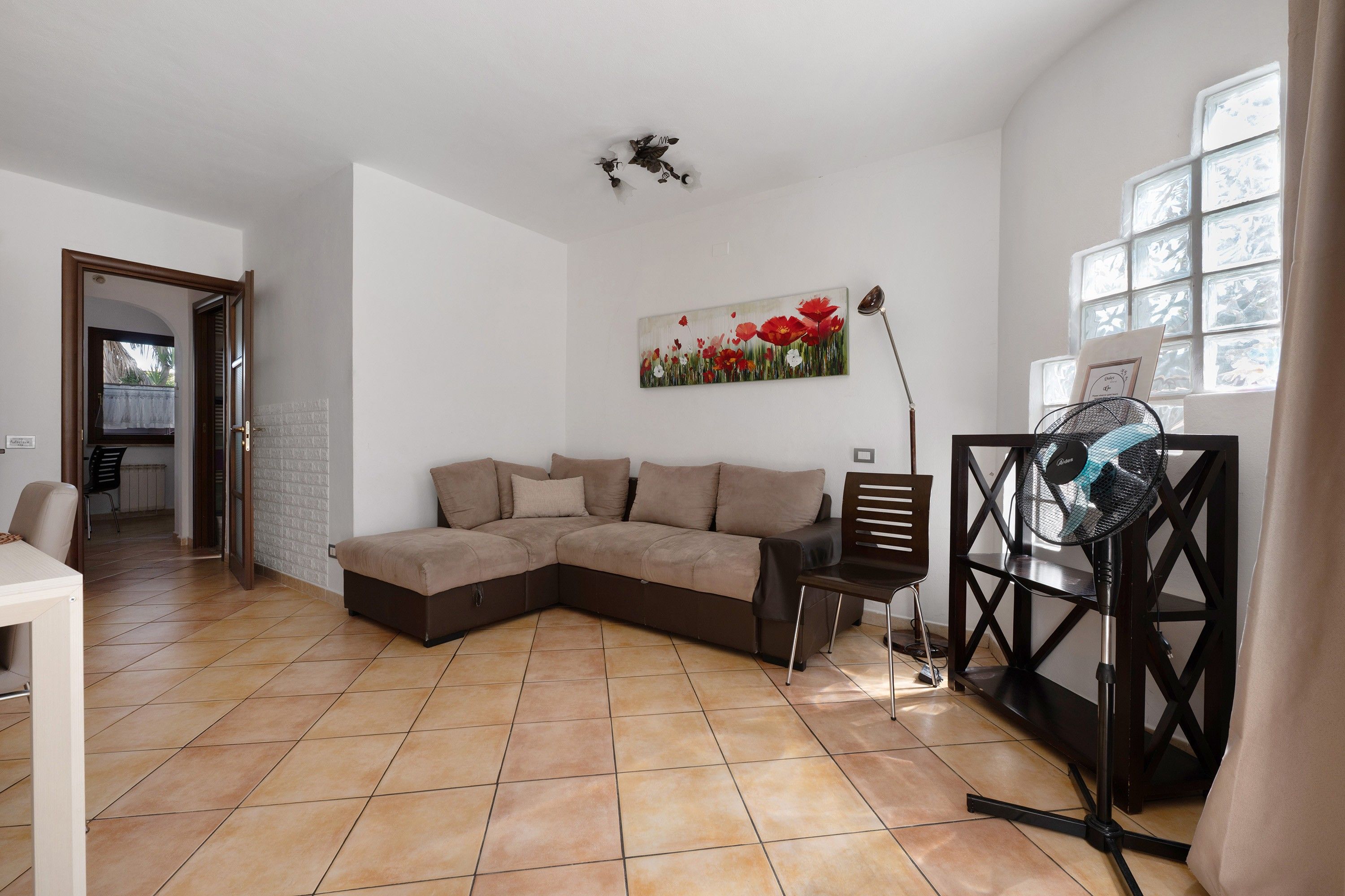 Ein Wohnzimmer mit Ecksofa, einem Blumenbild, einem Standventilator und einem Fenster mit Jalousien.
