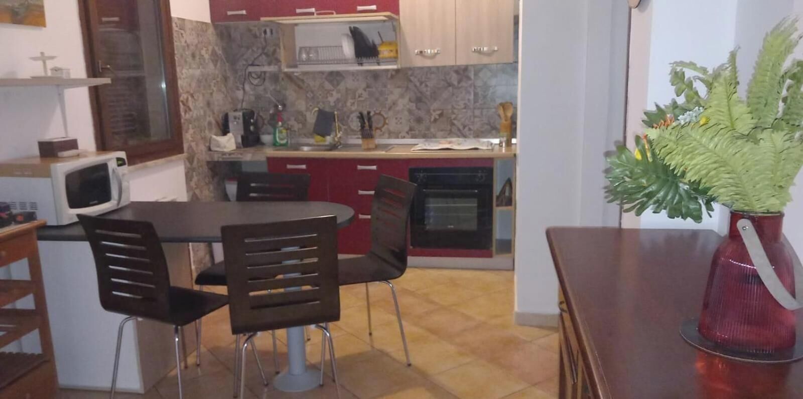 Une cuisine compacte équipée d’un four à micro-ondes, d’une cuisinière et de placards rouges. On y trouve une table à manger avec des chaises, et un pot de fleurs placé sur la droite.