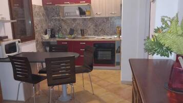 Une cuisine compacte équipée d’un four à micro-ondes, d’une cuisinière et de placards rouges. On y trouve une table à manger avec des chaises, et un pot de fleurs placé sur la droite.