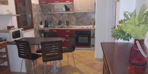 Una cucina compatta con forno a microonde, fornello e pensili rossi. Si nota un tavolo con sedie e una pianta in vaso alla destra.