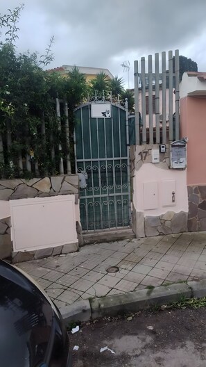 Ein verschlossenes Eingangstor mit einem Metallgitter, einer Steinmauer und einem befestigten Bereich, auf dem ein Auto teilweise sichtbar ist.