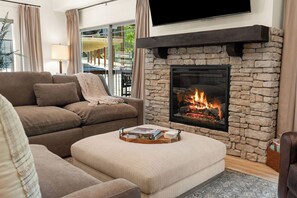 A living room with a stone fireplace, a sofa, a coffee table with books, and a TV mounted above the fireplace. - Wooded Cabin, Hot Tub | Pool, Fishing, Pickleball (Hollister)