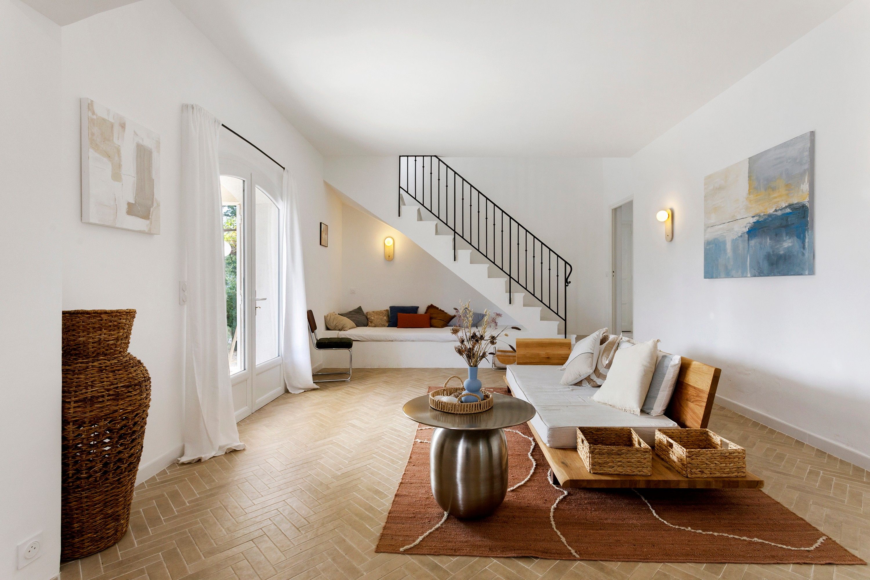 A modern living room with a herringbone floor, a staircase, a sofa set, and a large abstract painting.
