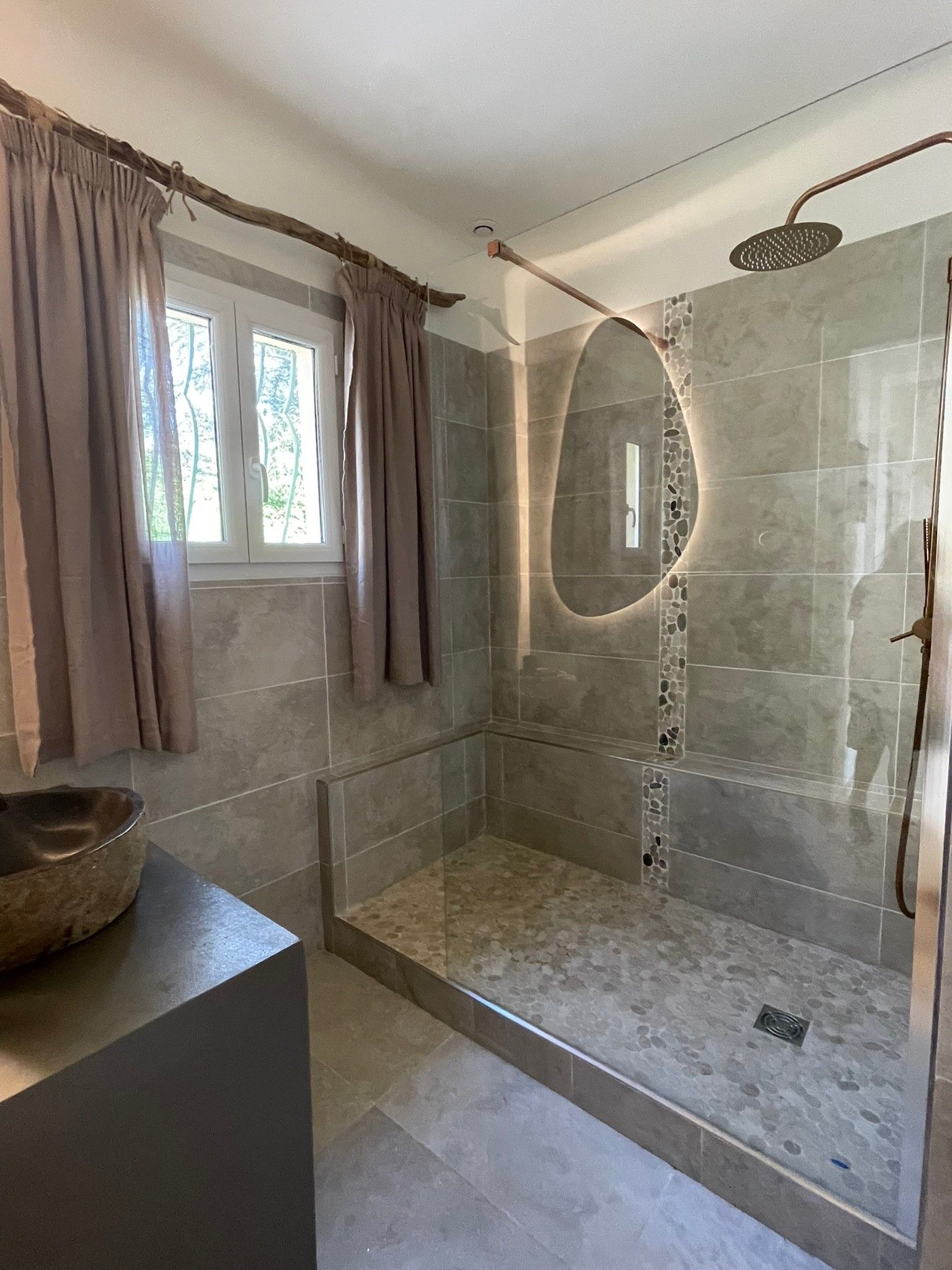 Une douche avec une porte en verre, un mur en pierre et une fenêtre avec des rideaux.