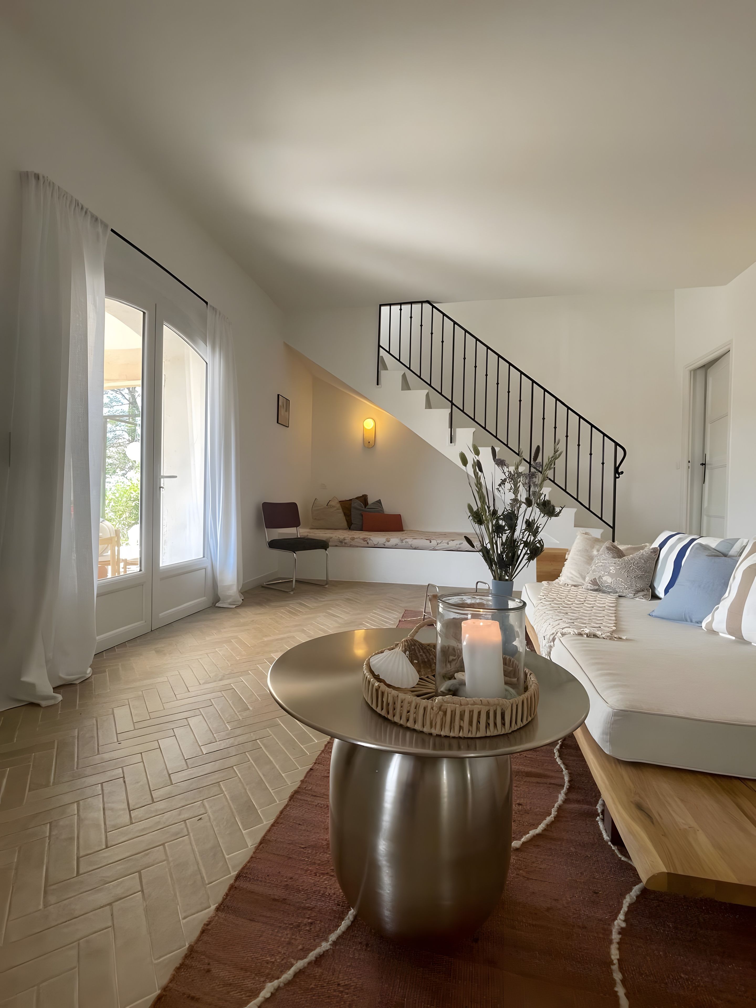 A modern living room with a herringbone floor, a round coffee table with candles, a sofa with patterned cushions, a bench with a cushion, and a staircase with a black railing.