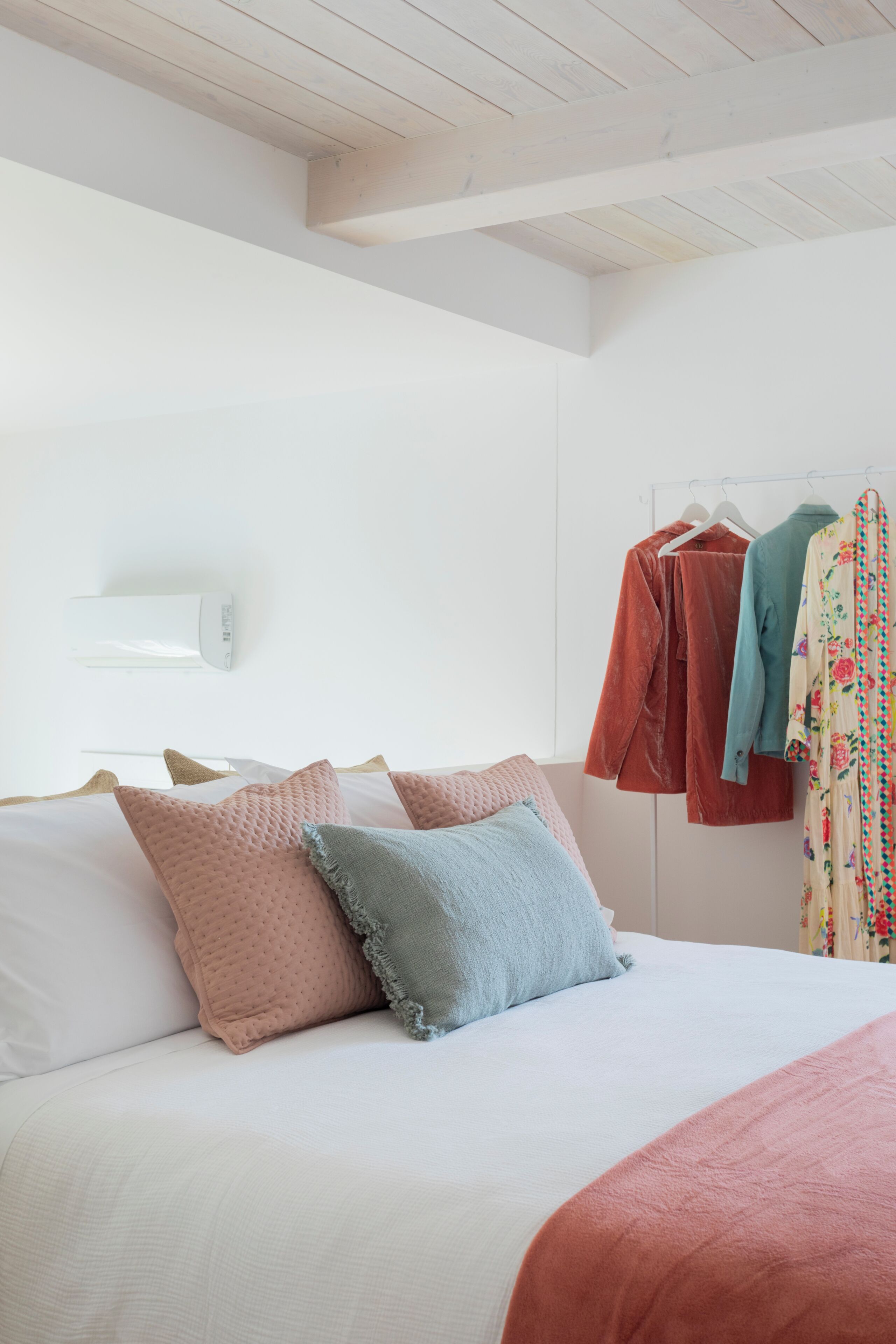 Ein Schlafzimmer mit einem Bett, Kissen, einer Pendelleuchte und einem Kleiderständer mit Kleidungsstücken.