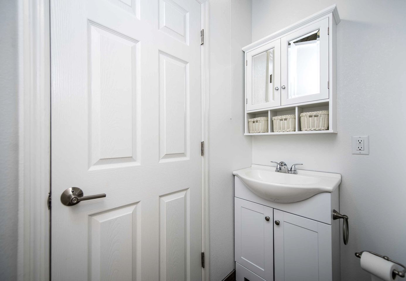 Un baño con puerta blanca, un lavabo, un espejo y una repisa con cestas.