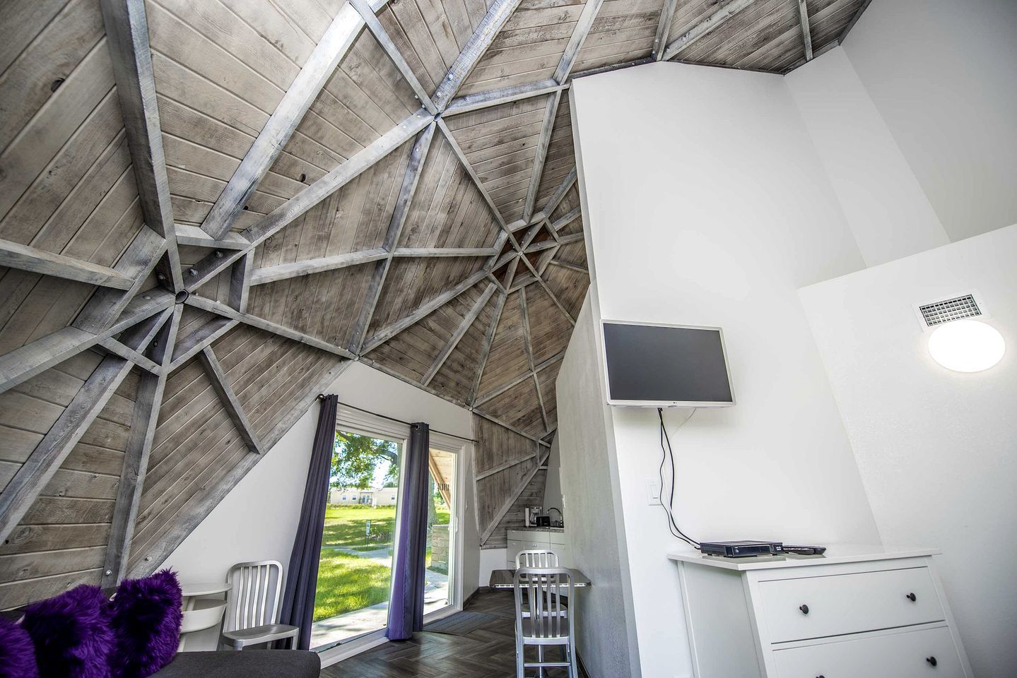 Une pièce avec un plafond en pente, un téléviseur fixé au mur, un bureau avec une chaise et une fenêtre avec des rideaux.