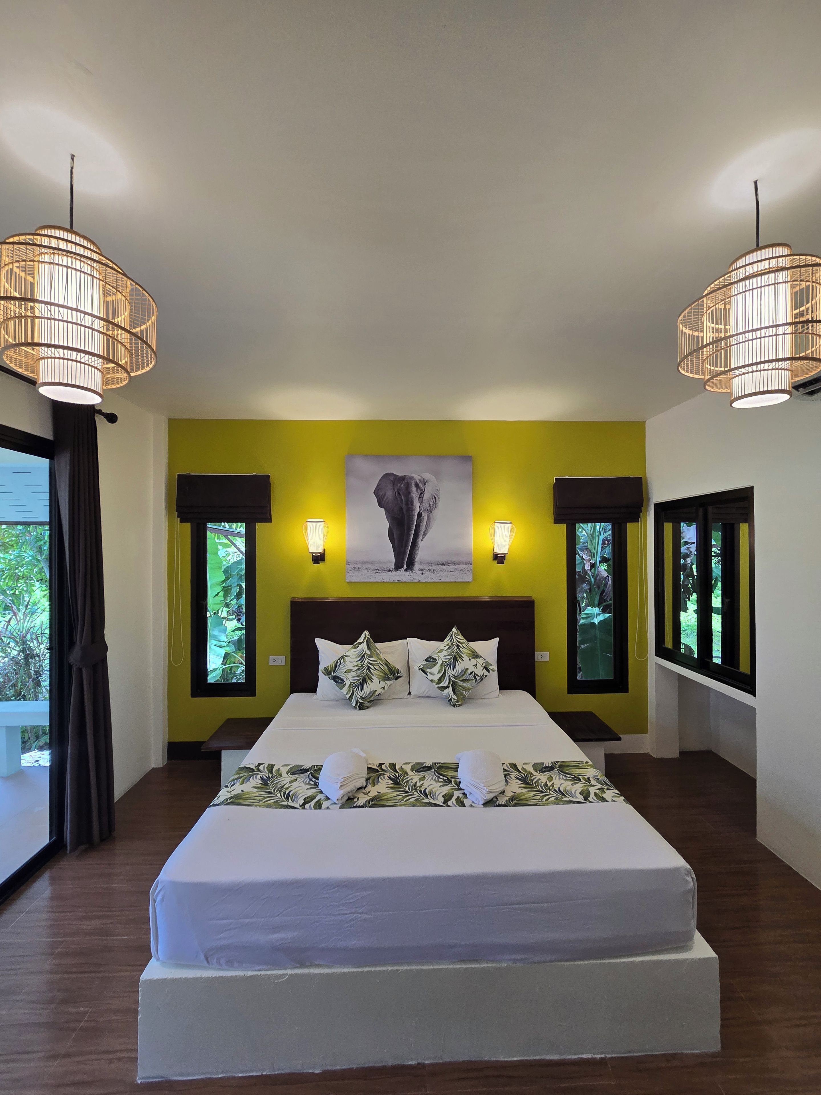 A hotel room with a large bed, two bedside lamps, a wall-mounted elephant picture, and a window with a view of greenery.