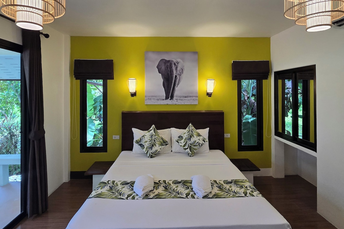 A hotel room with a large bed, two bedside lamps, a wall-mounted elephant picture, and a window with a view of greenery.