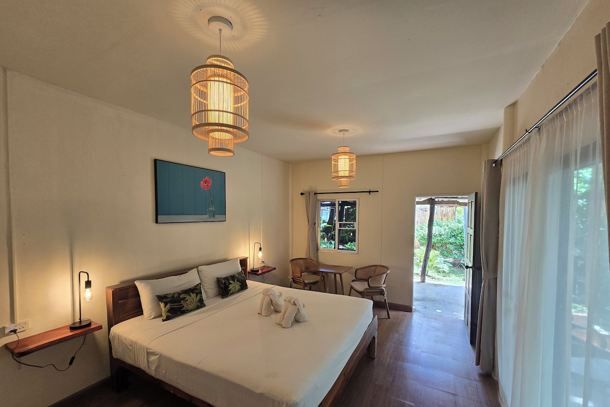 A hotel room with a large bed, bedside lamps, a dining area with chairs, and a view of greenery outside.