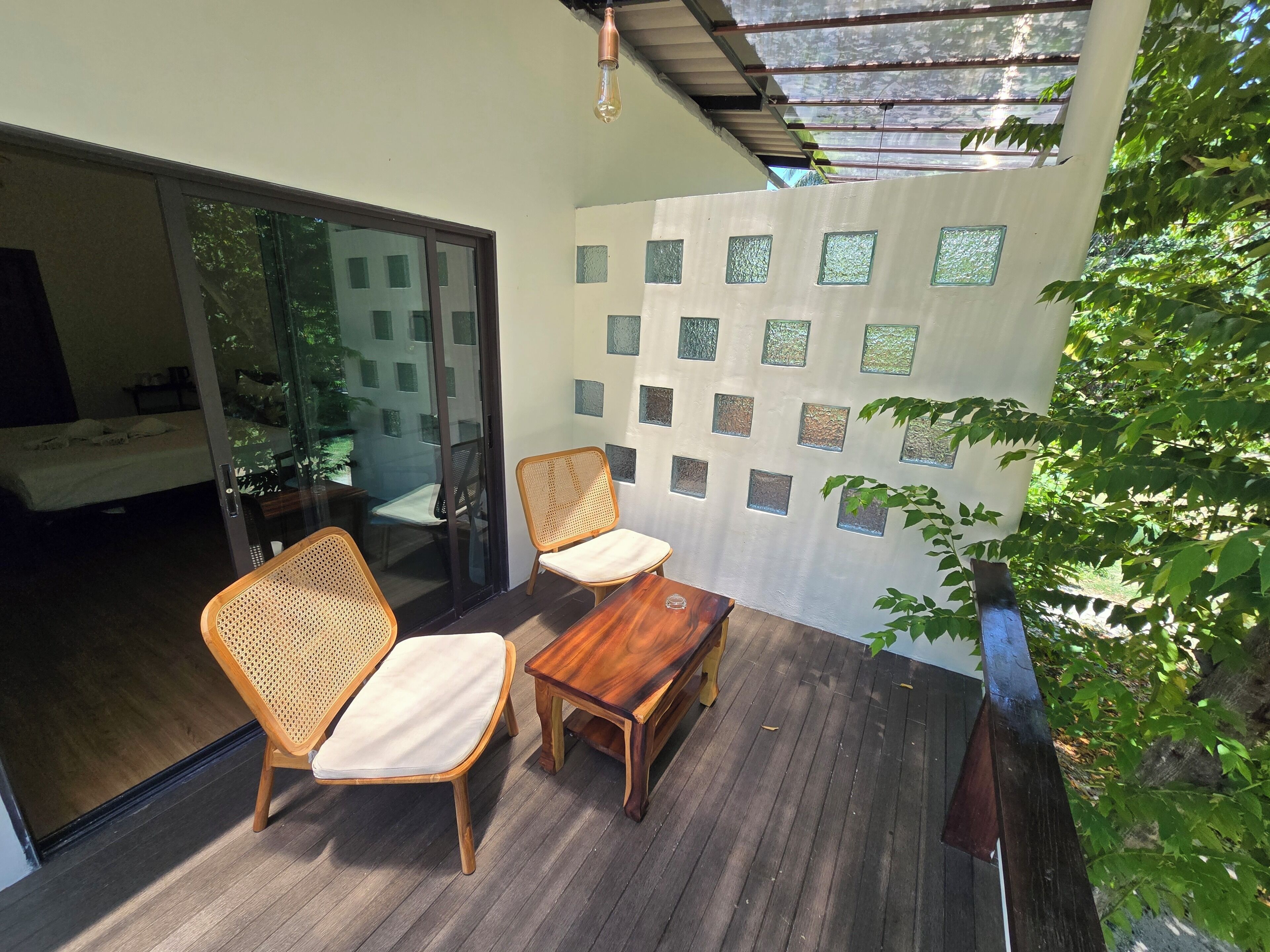 A wooden deck with two wicker chairs and a small table, a sliding glass door leading to a bedroom, and a wall with decorative square panels.
