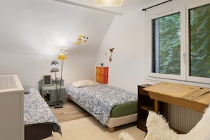 A bedroom with two beds, a desk, a lamp, and a window with a view of trees.