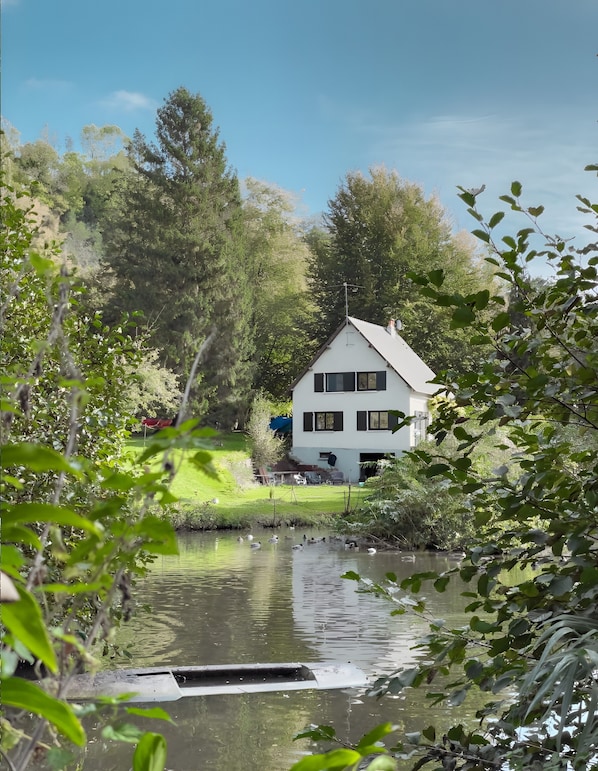 A house with a sloped roof, surrounded by trees and a body of water.