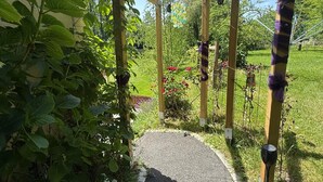 Puutarhapolku johtaa puiseen pergolaan, jonka yläpuolella on roikkuvia valaisimia. Polun ympärillä on vehreää kasvillisuutta ja kukkivia kasveja.