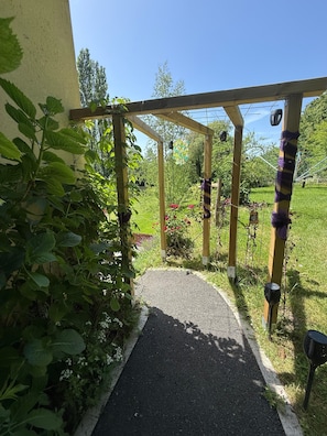 A garden pathway leading to a wooden pergola with hanging lights, surrounded by lush greenery and flowering plants.
