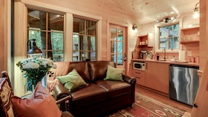 A cozy cabin interior with a leather sofa, a dining table with chairs, a kitchen area with a microwave and sink, and large windows offering a view of the outdoors.