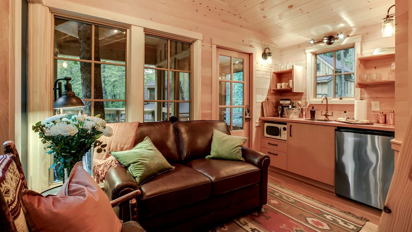 Un interior de cabaña acogedor con un sofá de cuero, una mesa de comedor con sillas, una zona de cocina con microondas y fregadero, y amplias ventanas con vistas al exterior.