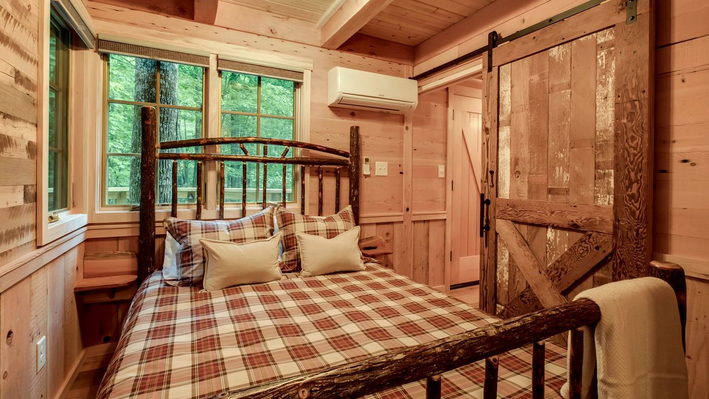 Een slaapkamer in een houten chalet, met een bed, kussens, een raam en een airconditioning.