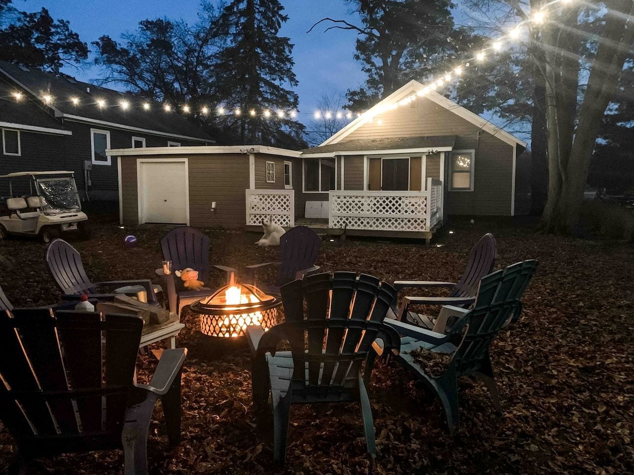 An outdoor seating area with chairs arranged around a fire pit, a covered porch, and string lights.