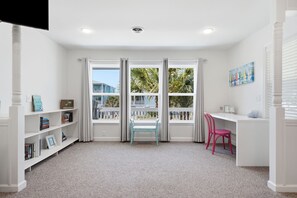 Ein Zimmer mit weißem Schreibtisch, Bücherregal, Stuhl und großen Fenstern mit Vorhängen.