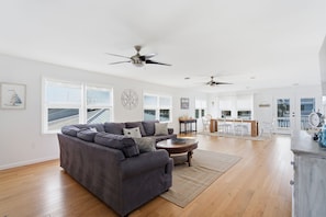 A spacious living room with a gray sectional sofa, a coffee table, and a ceiling fan. There are large windows and a view of the outside. - Fire Pit | Pet-Friendly | 2 Blocks to Pier (Kure Beach)