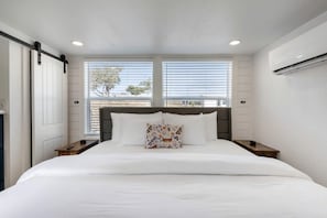 A bedroom with a bed, two bedside tables, a window with blinds, and an air conditioning unit. - Luxury Tiny Home Hideaway with Resort-Style Amenities Near Fredericksburg, TX (Fredericksburg)