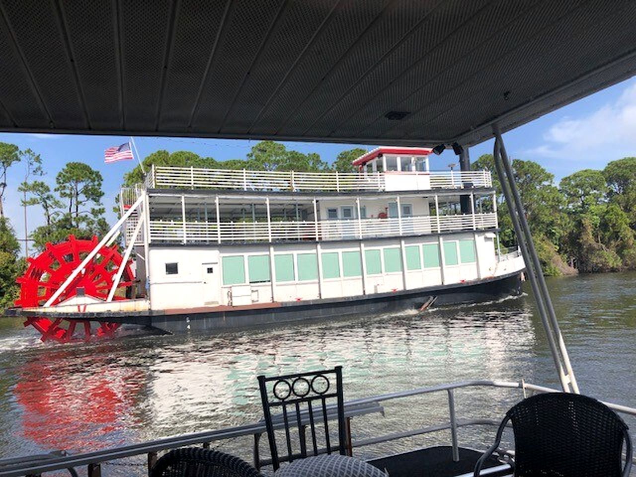 A riverboat with a large paddle wheel, a covered upper deck, and a flag.