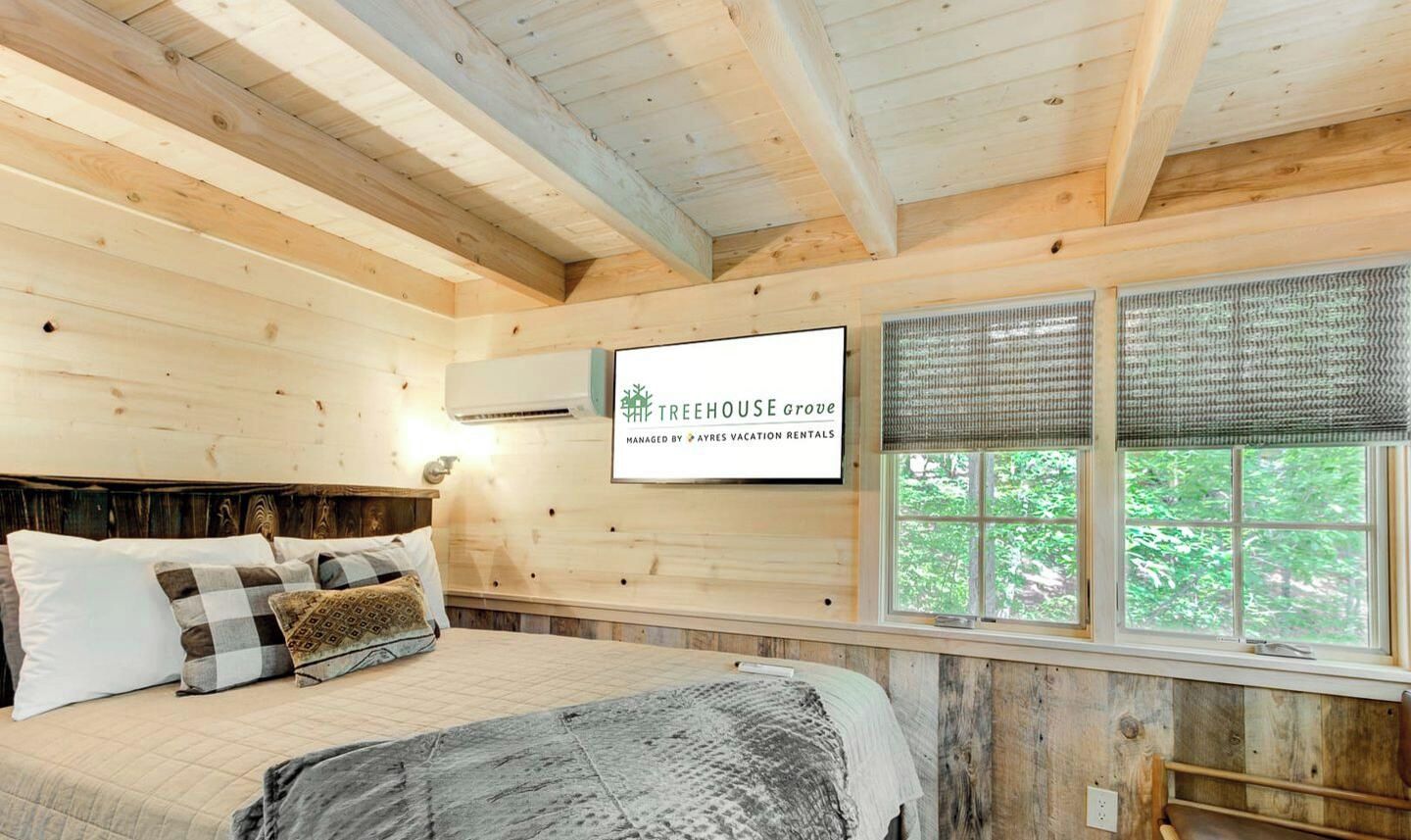 Een houten slaapkamer met een bed, kussens, een wandtelevisie en een raam met zonwering.