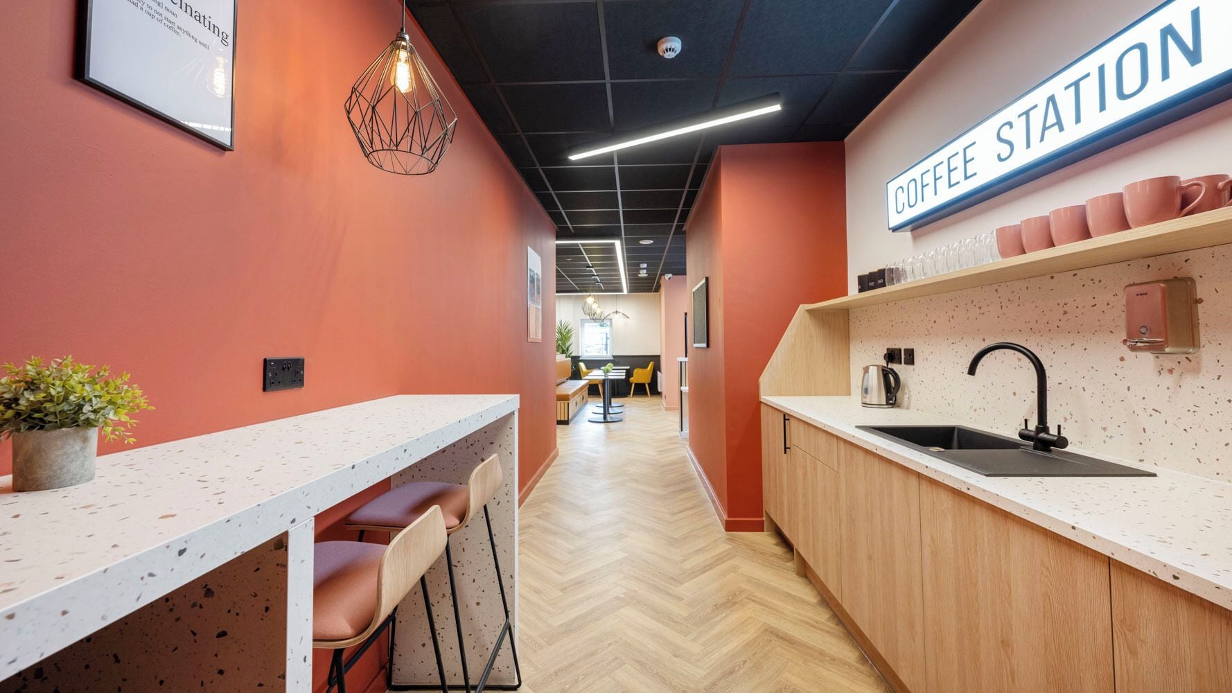 A modern kitchen area with a coffee station, bar seating, and a herringbone wood floor.