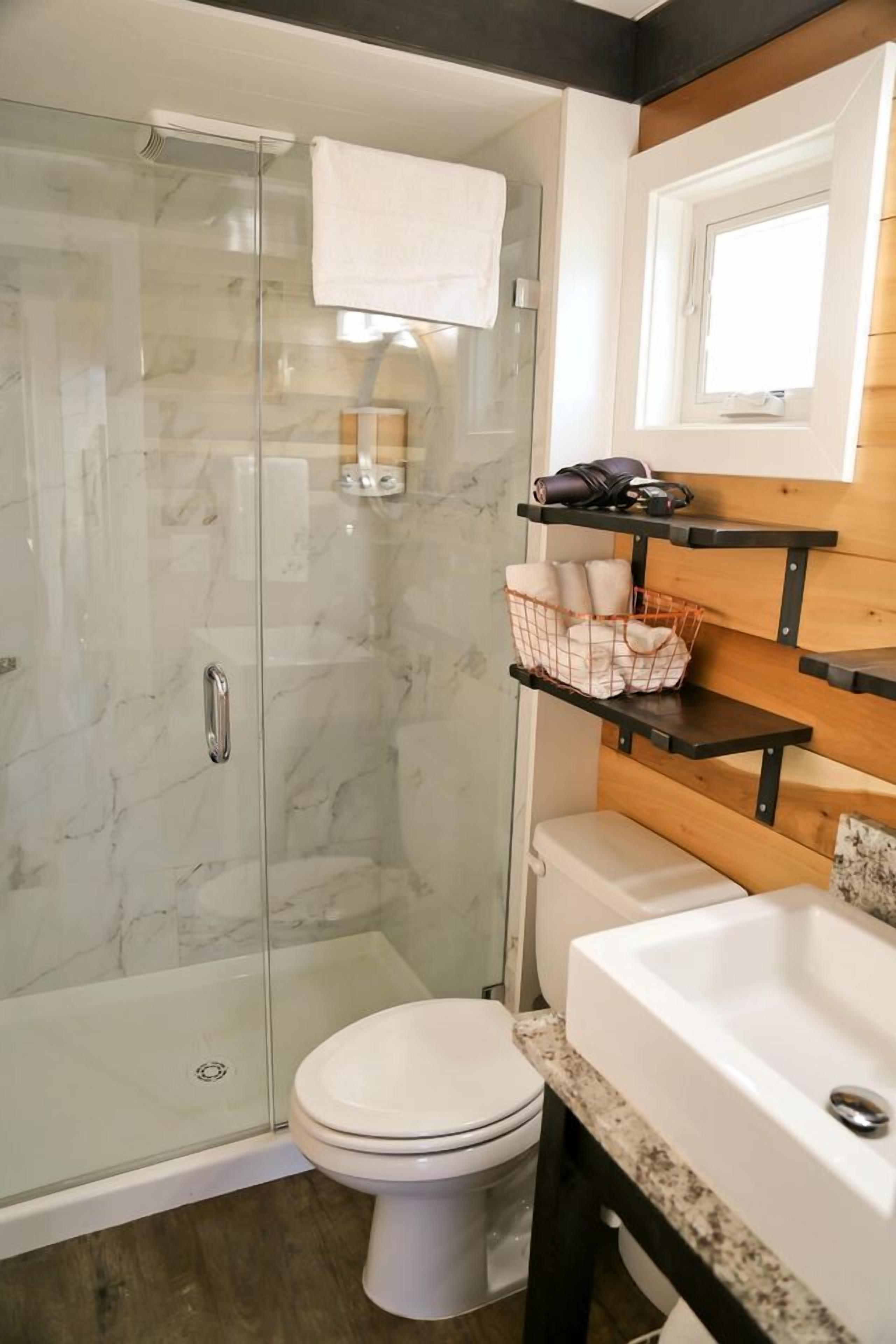 Een badkamer met een glazen douchecabine, een wit toilet, een wastafel, en houten planken met handdoeken en toiletartikelen.