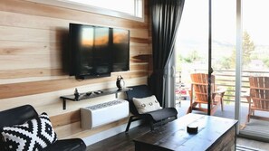 A modern living room with a flat-screen TV mounted on a wooden wall, a sofa with a patterned pillow, a coffee table, and a sliding glass door leading to an outdoor area with a chair.
