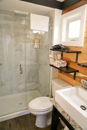 A bathroom with a glass-enclosed shower, a white toilet, a sink, and wooden shelves with towels and toiletries.