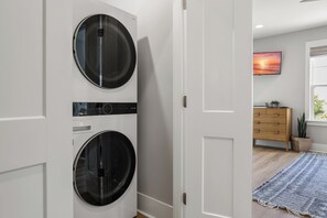 A laundry room with two front-loading washing machines, a wooden dresser, a painting of a sunset, and a blue rug on the floor. - 4BR Beach Home | Pet Friendly | Walk to Ocean (Kure Beach)