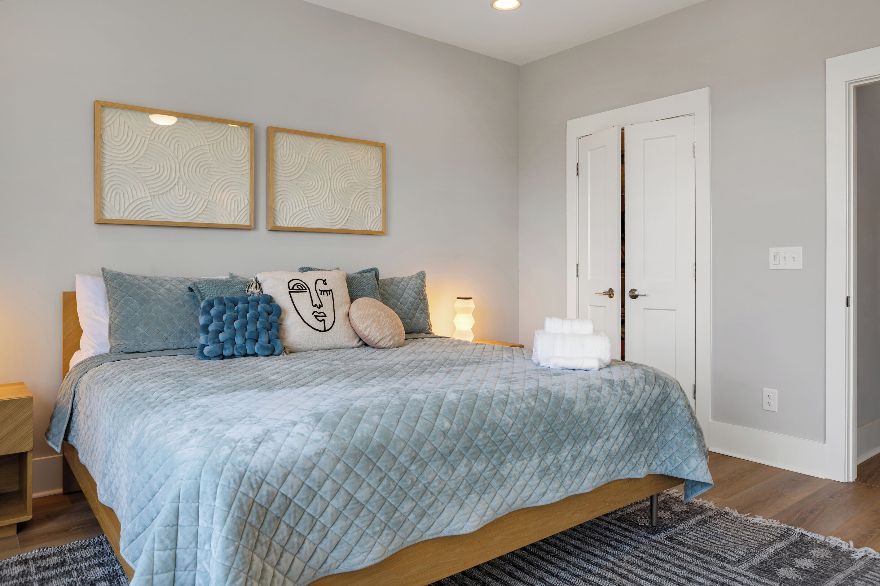 A bedroom with a bed, pillows, a bedside table, a nightstand, a wall-mounted light, a door, and a rug.