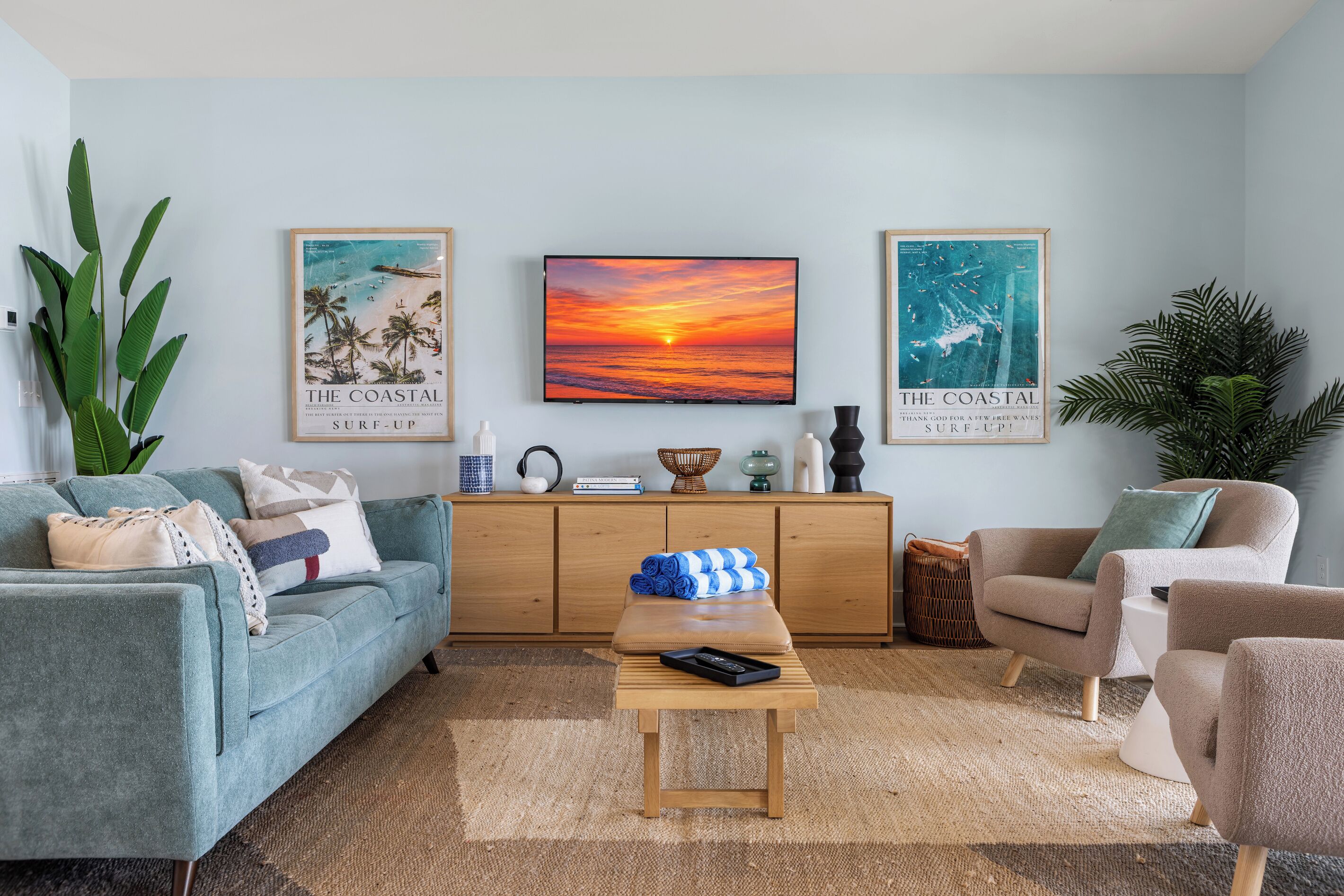 A living room with a sofa, armchair, coffee table, and a TV on the wall. There are two posters on the wall, a plant, and a cabinet with items on top.