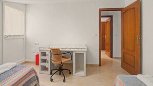 Ein Zimmer mit Deckenventilator, Schreibtisch mit Stuhl, Fenster mit Jalousien und einer Tür zu einem weiteren Raum.