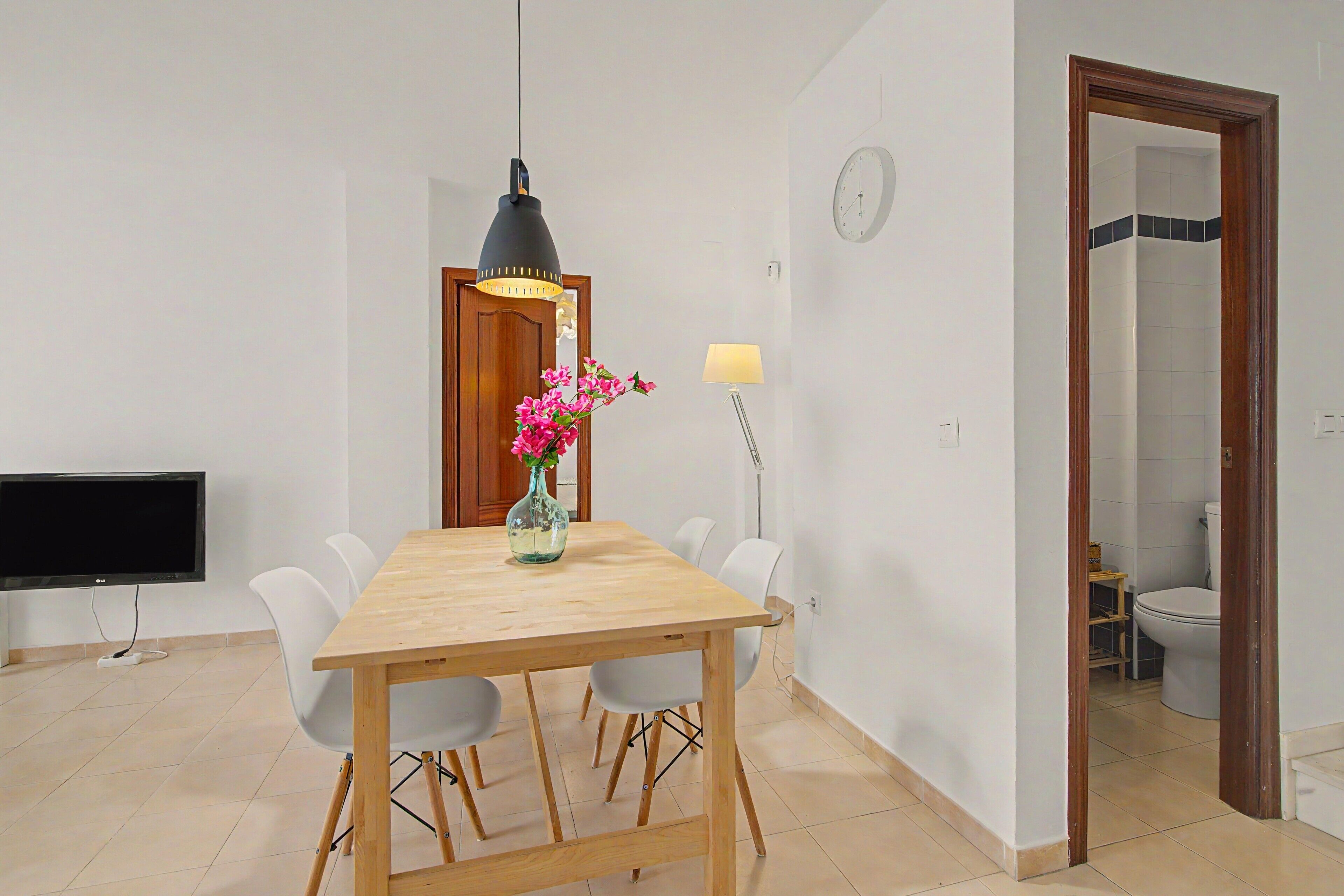 Un comedor con una mesa de madera, sillas, un televisor, un florero con flores, una lámpara y un reloj en la pared.