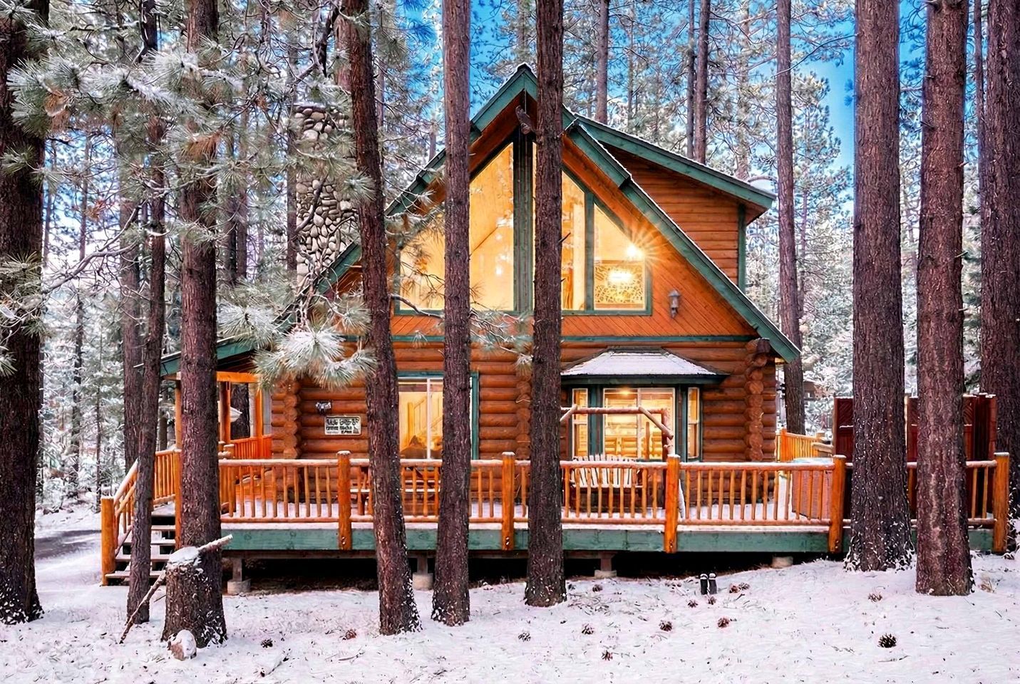 A log cabin with a wooden deck and a covered porch, surrounded by tall pine trees in a snowy forest.