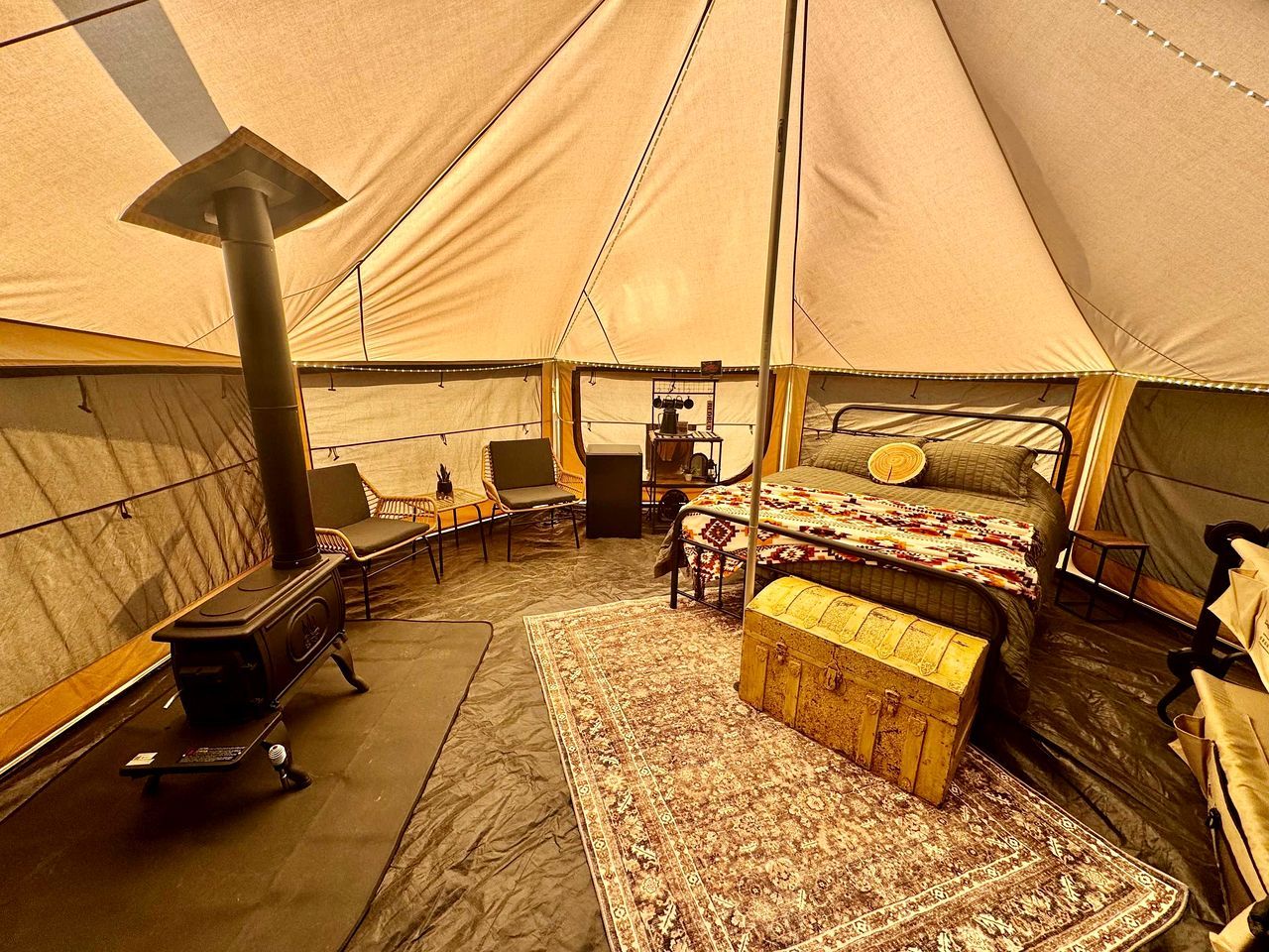 A tent interior with a bed, seating area, and a wood-burning stove.