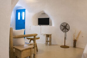Ein Zimmer mit einem Holzstuhl, einem kleinen Tisch, einem Standventilator und einem an der Wand befestigten Fernseher.