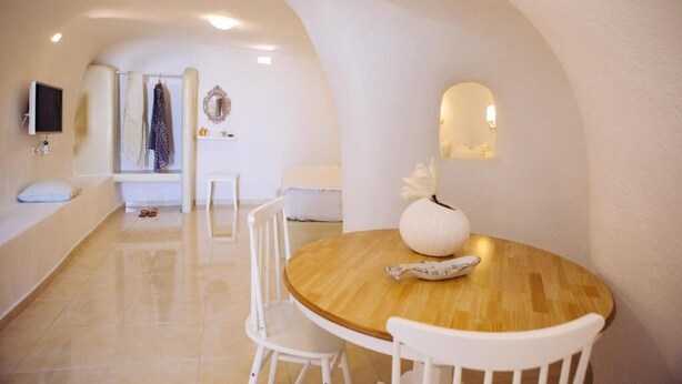 Um interior moderno com uma mesa de madeira redonda, cadeiras brancas, uma televisão fixada na parede e uma parede branca com um espelho decorativo.