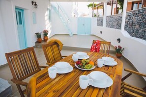 Een houten eettafel gedekt met borden, kopjes en een fruitschaal in een zonnig buitengebied, met een blauwe deur en een stenen muur.